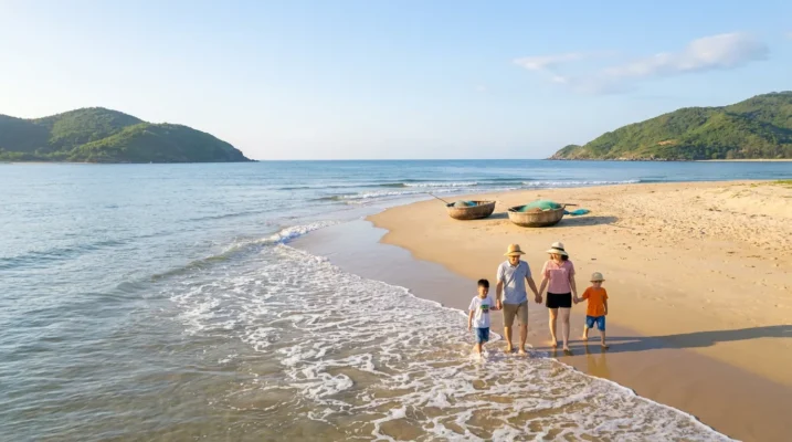 Bãi biển hình cánh cung ở Bắc Trung Bộ buổi sáng mùa hè, hai ngọn núi thấp ôm hai đầu bãi, người Việt dạo trên cát và thuyền thúng gần bờ — ảnh minh họa